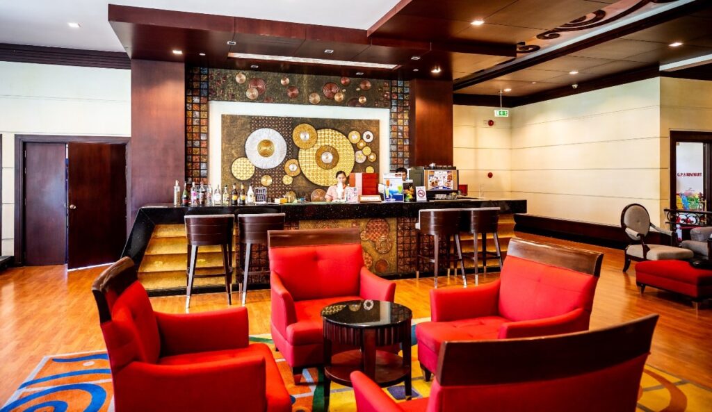 The Grand Pacific Sovereign Resort & Spa lobby features a reception desk with a vibrant, circular patterned backdrop. Red armchairs and small black tables are arranged on a hardwood floor. The reception area includes a bar counter with high stools, and a staff member is present behind the desk.