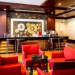 The Grand Pacific Sovereign Resort & Spa lobby features a reception desk with a vibrant, circular patterned backdrop. Red armchairs and small black tables are arranged on a hardwood floor. The reception area includes a bar counter with high stools, and a staff member is present behind the desk.