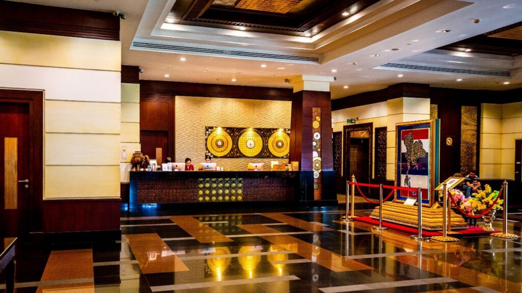 The Grand Pacific Sovereign Resort & Spa lobby features a polished floor, a check-in counter with attentive staff, and decorative wall accents. A roped-off display area to the right showcases a decorative piece and a floral arrangement. Warm lighting sets a cozy ambiance, enhancing this prime hotel investment opportunity.