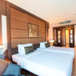 A hotel room at the Grand Pacific Sovereign Resort & Spa with two neatly made single beds, each with white linens and green accent pillows. The room features wooden furnishings, a plush padded headboard, and soft lighting from bedside lamps. Double doors open to a well-lit area with additional decor.