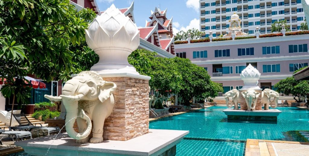 A serene outdoor pool area with ornate elephant statues holding rings at their trunks on large pedestals. The pool is surrounded by lush greenery and has a backdrop of traditional Thai-style buildings with red roofs and a modern multi-story building, embodying the charm of Grand Pacific Sovereign Resort & Spa.