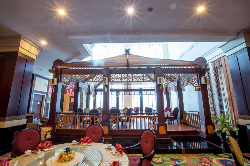 The image shows an interior of a restaurant within the Grand Pacific Sovereign Resort & Spa, featuring a wooden gazebo structure in the center. Elegant round tables with white tablecloths, red napkins, and patterned carpet adorn the space, while warm lighting creates a cozy and welcoming atmosphere.