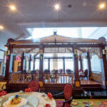 The image shows an interior of a restaurant within the Grand Pacific Sovereign Resort & Spa, featuring a wooden gazebo structure in the center. Elegant round tables with white tablecloths, red napkins, and patterned carpet adorn the space, while warm lighting creates a cozy and welcoming atmosphere.