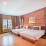 A spacious room at the Grand Pacific Sovereign Resort & Spa features two neatly made double beds, a large window with beige curtains offering a view of trees and rooftops, wooden flooring, a flat-screen TV on a dark wood dresser, and modern decor in brown and beige tones.