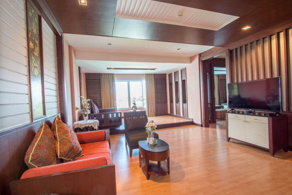 A spacious, well-lit living room with wooden flooring and furnishings. It features a red sofa with decorative pillows, a round coffee table, and a large flat-screen TV on a white console. Large windows with curtains offer views of the Grand Pacific Sovereign Resort & Spa and natural light fills the room.