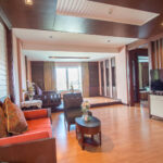 A spacious, well-lit living room with wooden flooring and furnishings. It features a red sofa with decorative pillows, a round coffee table, and a large flat-screen TV on a white console. Large windows with curtains offer views of the Grand Pacific Sovereign Resort & Spa and natural light fills the room.
