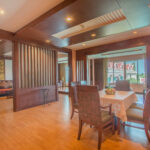 A spacious room with wooden flooring and modern wooden paneling features a dining table with four upholstered chairs. A small centerpiece adorns the table. In the background, a cozy living area with a couch and two outdoor balconies can be seen, reminiscent of the elegance found at Grand Pacific Sovereign Resort & Spa.