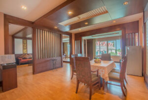 A spacious room with wooden flooring and modern wooden paneling features a dining table with four upholstered chairs. A small centerpiece adorns the table. In the background, a cozy living area with a couch and two outdoor balconies can be seen, reminiscent of the elegance found at Grand Pacific Sovereign Resort & Spa.