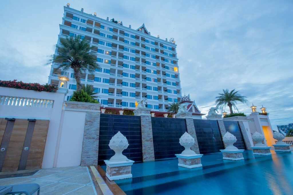 A modern, multi-story hotel with multiple balconies is seen from a low angle at dusk. The Grand Pacific Sovereign Resort & Spa is flanked with palm trees and features an elaborate pool area in the foreground. The sky is cloudy, giving a serene ambiance to this off-market hotel investment opportunity.