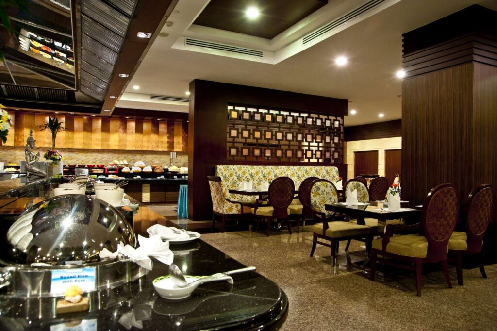 A restaurant interior at the Grand Pacific Sovereign Resort & Spa features a buffet setup on the left side with a variety of dishes. The seating area on the right offers cushioned chairs and wooden tables. Dark wood accents and modern lighting adorn the space, while the polished floor adds a reflective touch.
