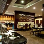 A restaurant interior at the Grand Pacific Sovereign Resort & Spa features a buffet setup on the left side with a variety of dishes. The seating area on the right offers cushioned chairs and wooden tables. Dark wood accents and modern lighting adorn the space, while the polished floor adds a reflective touch.