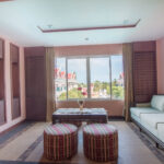 A bright living room with peach-colored walls, a large square window offering a view of buildings and trees, a white cushioned sofa, two round ottomans with striped patterns, and a rectangular coffee table with decorative items; reminiscent of the luxurious ambiance you'd find at the Grand Pacific Sovereign Resort & Spa.