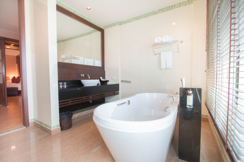 A modern bathroom at the Grand Pacific Sovereign Resort & Spa features a large white freestanding bathtub in the center, a mounted black hand shower next to it, twin sinks with a dark wooden shelf underneath, wide mirror, towel rack with rolled towels, and large window blinds letting in natural light and fresh sea breezes.