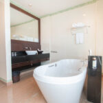 A modern bathroom at the Grand Pacific Sovereign Resort & Spa features a large white freestanding bathtub in the center, a mounted black hand shower next to it, twin sinks with a dark wooden shelf underneath, wide mirror, towel rack with rolled towels, and large window blinds letting in natural light and fresh sea breezes.