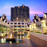 A lavish resort with traditional architectural elements is shown at dusk. The Grand Pacific Sovereign Resort & Spa features a central courtyard with illuminated fountains, surrounded by elegant buildings with intricate roof designs. A tall building is visible in the background against a purple sky.
