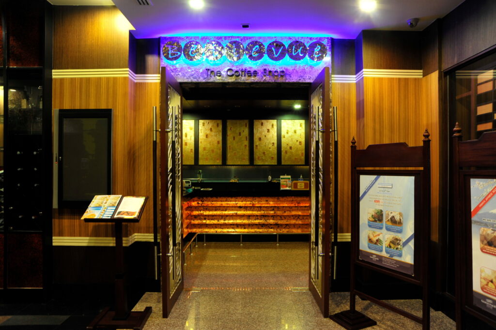 Entrance of a coffee shop with an illuminated sign displaying "Bayswater The Coffee Shop." Two wooden display stands with menus flank the doorway. The interior features warm lighting, wooden finishes, and a counter at the back, reminiscent of the cozy charm you'd find at the Grand Pacific Sovereign Resort & Spa.
