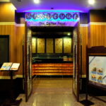 Entrance of a coffee shop with an illuminated sign displaying "Bayswater The Coffee Shop." Two wooden display stands with menus flank the doorway. The interior features warm lighting, wooden finishes, and a counter at the back, reminiscent of the cozy charm you'd find at the Grand Pacific Sovereign Resort & Spa.