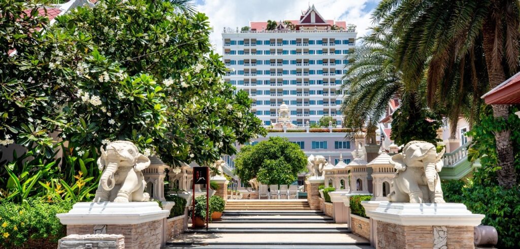 An exterior view of the Grand Pacific Sovereign Resort & Spa reveals a white, multi-story building in the background. The pathway to the hotel entrance is lined with trees, greenery, and stone statues of elephants on both sides. Palm trees and manicured plants enhance the tropical atmosphere.
