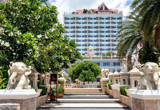 An exterior view of the Grand Pacific Sovereign Resort & Spa reveals a white, multi-story building in the background. The pathway to the hotel entrance is lined with trees, greenery, and stone statues of elephants on both sides. Palm trees and manicured plants enhance the tropical atmosphere.