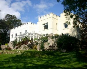 A large white hotel with a castle-like design, featuring battlements on the roof. The commercial building boasts numerous windows and a glass conservatory. Surrounded by lush greenery, rocks, and trees, with a well-kept lawn in the foreground, it offers significant venue and development potential.