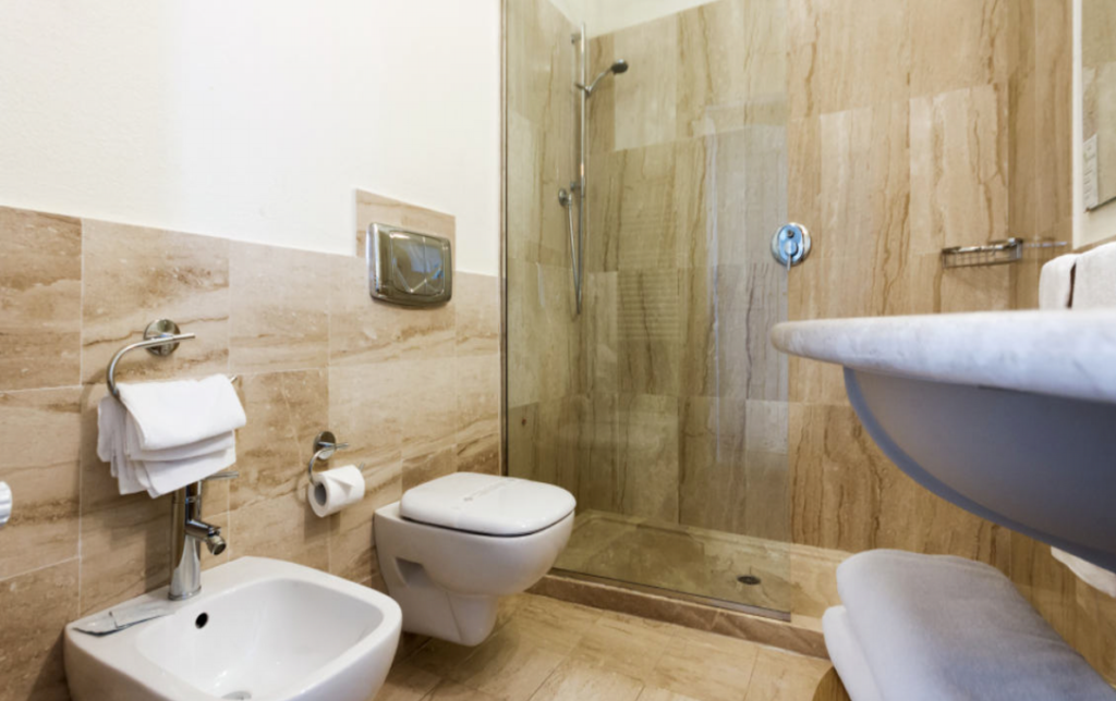 A modern bathroom with beige marble tiles, featuring a glass-enclosed shower, a wall-mounted toilet, and a bidet. A white towel hangs neatly on a ring near the bidet and a toilet paper roll is placed beside the toilet. A curved sink is visible on the right.