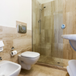A modern bathroom with beige marble tiles, featuring a glass-enclosed shower, a wall-mounted toilet, and a bidet. A white towel hangs neatly on a ring near the bidet and a toilet paper roll is placed beside the toilet. A curved sink is visible on the right.