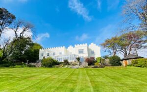 Property for sale by Auction A spacious, well-manicured lawn leads up to Nicholaston House in Swansea, a large, white, castle-like building with crenellations. This hotel and commercial building with significant venue and development potential features a sunroom with large windows surrounded by lush greenery and tall trees against a vibrant blue sky. Presented by Palace Auctions