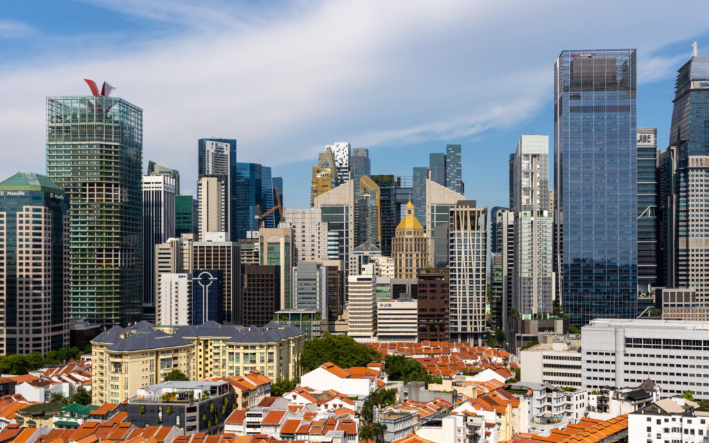 A panoramic view of a city skyline in Singapore features a mix of modern skyscrapers and older, lower-rise buildings with red-tiled roofs in the foreground. The sky is mostly clear with a few light clouds, and the tallest glass-and-steel structures hint at lucrative office investment opportunities.