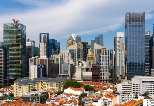 A panoramic view of a city skyline in Singapore features a mix of modern skyscrapers and older, lower-rise buildings with red-tiled roofs in the foreground. The sky is mostly clear with a few light clouds, and the tallest glass-and-steel structures hint at lucrative office investment opportunities.