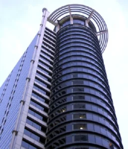 An upward view of a modern, cylindrical skyscraper with reflective glass windows and a unique circular rooftop structure with vertical bars. Adjacent to a rectangular, multi-story structure with a patterned facade, this prime piece of office investment in Singapore is showcased against a clear sky.