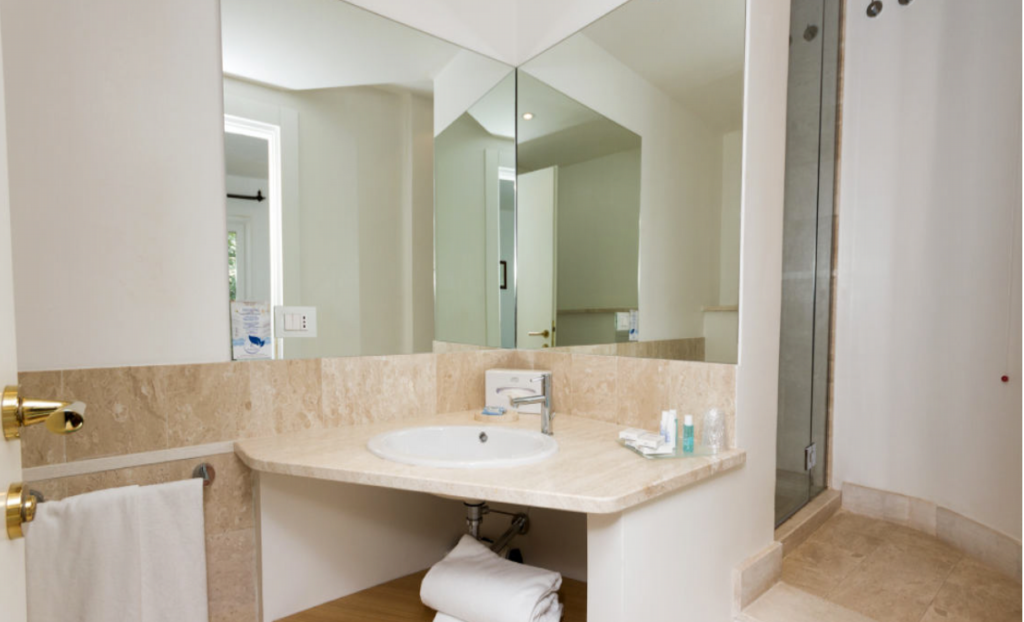 A modern bathroom features a large mirror, a beige marble countertop with a white sink, and well-organized toiletries including a glass, soap dispenser, and towels. To the right, there's a glass-enclosed shower with a tiled interior.