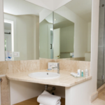 A modern bathroom features a large mirror, a beige marble countertop with a white sink, and well-organized toiletries including a glass, soap dispenser, and towels. To the right, there's a glass-enclosed shower with a tiled interior.