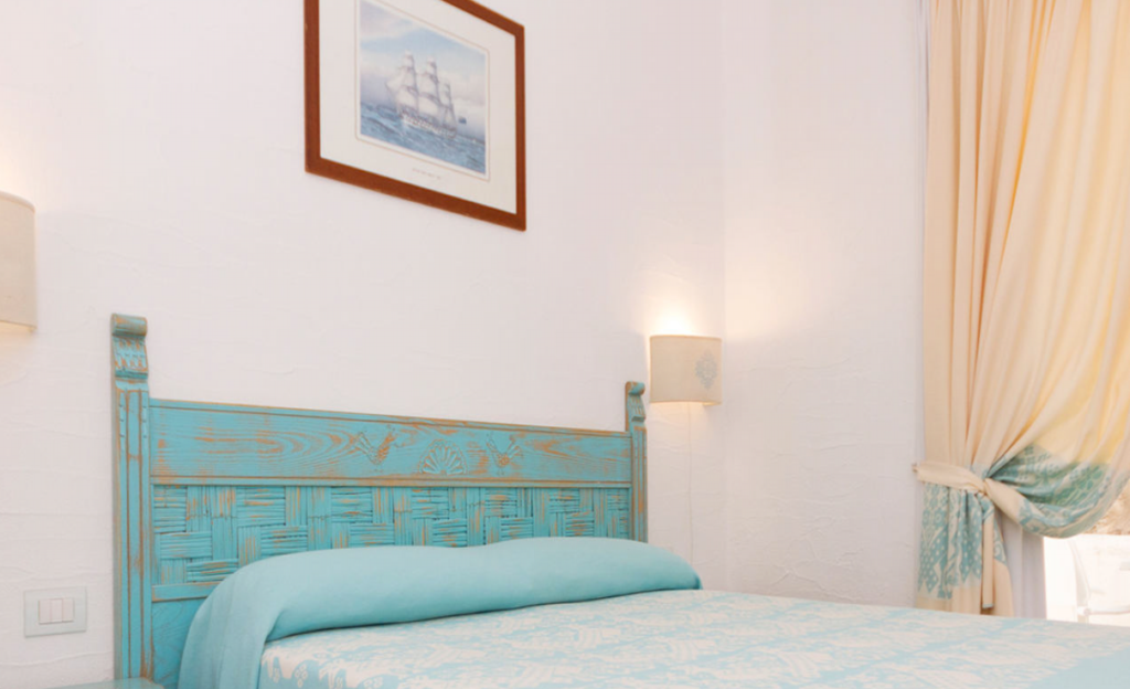 A cozy bedroom with a light teal ornate wooden headboard and a matching teal bedspread. The room features white walls with subtle texture, two wall-mounted reading lamps, and a framed nautical-themed picture above the bed. A curtain is tied back next to a window.