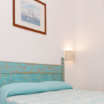 A cozy bedroom with a light teal ornate wooden headboard and a matching teal bedspread. The room features white walls with subtle texture, two wall-mounted reading lamps, and a framed nautical-themed picture above the bed. A curtain is tied back next to a window.