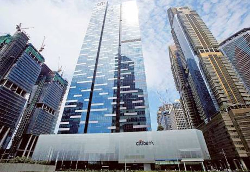 A modern urban scene featuring a high-rise building with a "Citibank" logo at the bottom. Flanked by other skyscrapers under construction, the blue sky with scattered clouds forms a backdrop. The main building, ideal for office investment business for sale in Singapore, has a sleek glass facade with geometric patterns.