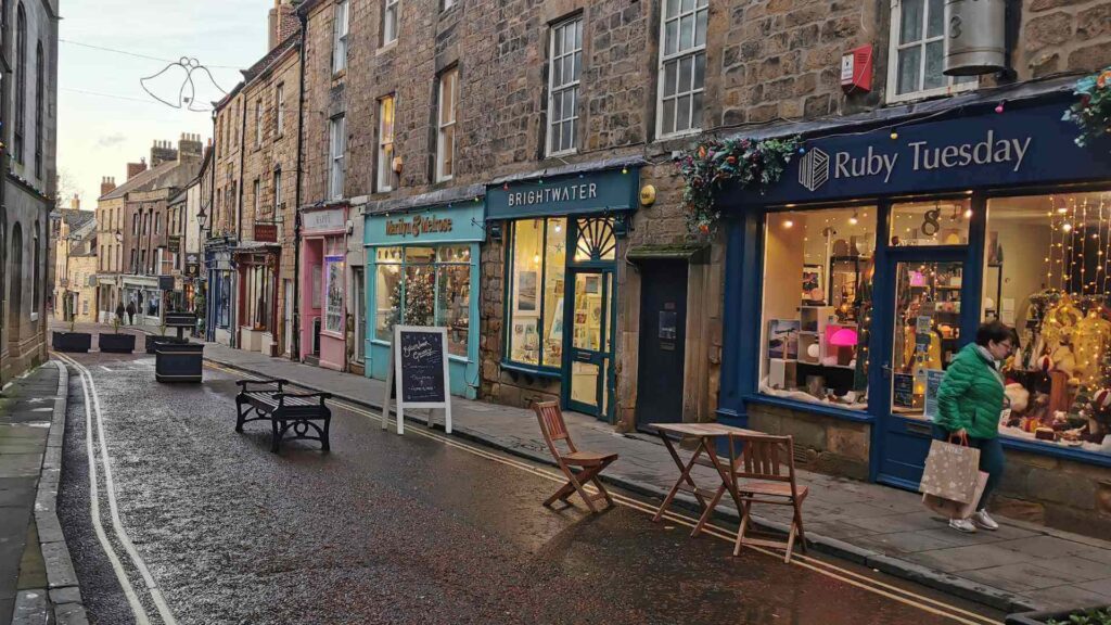 A quaint street lined with small shops, including "Bright Water" and "Ruby Tuesday." The brick buildings have large windows displaying goods. There are wooden tables and chairs on the sidewalk, and a person carrying a shopping bag strolls by. The street is wet, suggesting recent rain.