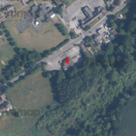 Aerial view of a residential area with multiple houses, roads, and patches of greenery. A red marker pinpoints a specific location near the edge of a forested area and an open field, close to the Three Lions Public house. The surrounding environment includes dense tree cover and nearby farmland.