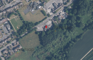 Aerial view of a residential area with multiple houses, roads, and patches of greenery. A red marker pinpoints a specific location near the edge of a forested area and an open field, close to the Three Lions Public house. The surrounding environment includes dense tree cover and nearby farmland.