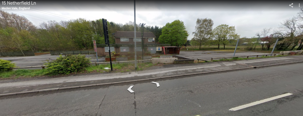 Street view of the closed Three Lions Public House, featuring a fenced outdoor area and a large parking lot. Situated on a roadside with nearby trees and greenery, the establishment's signage and building exhibit wear and disuse. Roadside arrows indicate it is a street view map of this potential development site.