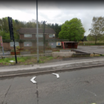 Street view of the closed Three Lions Public House, featuring a fenced outdoor area and a large parking lot. Situated on a roadside with nearby trees and greenery, the establishment's signage and building exhibit wear and disuse. Roadside arrows indicate it is a street view map of this potential development site.