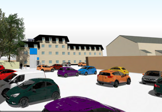 An illustrated image of a parking lot with various colored cars, including red, white, green, purple, orange, and blue. The lot is adjacent to a multi-story beige building with a dark roof and a nearby tree on the left. Other buildings are visible in the background.