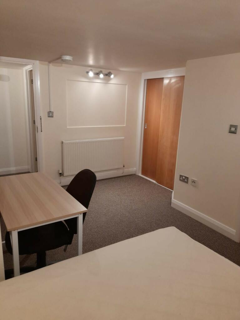 Property for sale by Auction A small, well-lit room with beige walls and carpeted floor. It features a wooden desk with a black chair, a bed partially visible in the foreground, a radiator on the back wall, a door on the right, and a three-bulb spotlight on the ceiling. Presented by Palace Auctions