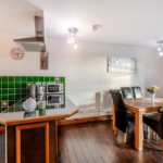 Property for sale by Auction A modern kitchen and dining area with wooden flooring at The Old Drapery Apartments, 12 Main St, Haltwhistle, for sale by auction. The kitchen features a green tiled backsplash, white cabinets, a microwave, and a kettle. An island counter separates it from the dining space, which has a wooden table with black leather chairs and a vase of flowers. Presented by Palace Auctions
