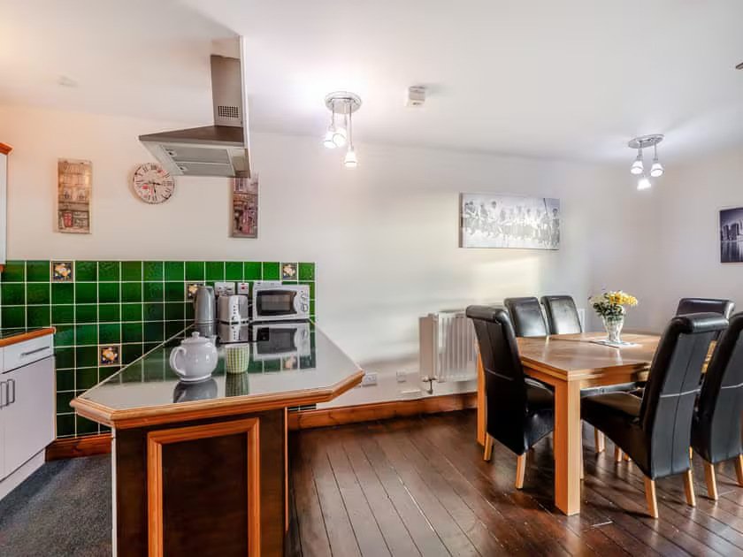 Property for sale by Auction A modern kitchen and dining area with wooden flooring at The Old Drapery Apartments, 12 Main St, Haltwhistle, for sale by auction. The kitchen features a green tiled backsplash, white cabinets, a microwave, and a kettle. An island counter separates it from the dining space, which has a wooden table with black leather chairs and a vase of flowers. Presented by Palace Auctions