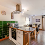 Property for sale by Auction A modern kitchen and dining area with wooden flooring at The Old Drapery Apartments, 12 Main St Haltwhistle, for sale by auction. The kitchen boasts a green tiled backsplash, white cabinets, and a central island with a stovetop and range hood. A wooden table set for six graces the dining area, complemented by artwork and a fan. Presented by Palace Auctions