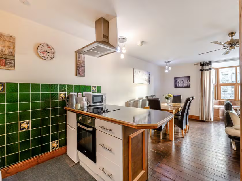 Property for sale by Auction A modern kitchen and dining area with wooden flooring at The Old Drapery Apartments, 12 Main St Haltwhistle, for sale by auction. The kitchen boasts a green tiled backsplash, white cabinets, and a central island with a stovetop and range hood. A wooden table set for six graces the dining area, complemented by artwork and a fan. Presented by Palace Auctions