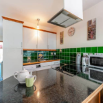 Property for sale by Auction A modern kitchen with green tiled backsplash and white cabinets in The Old Drapery Apartments at 12 Main St Haltwhistle, for sale by auction. The countertop features a teapot, a cup, and a kettle. Appliances such as a microwave, coffeemaker, and oven are visible. A clock and paintings adorn the well-lit space. Presented by Palace Auctions