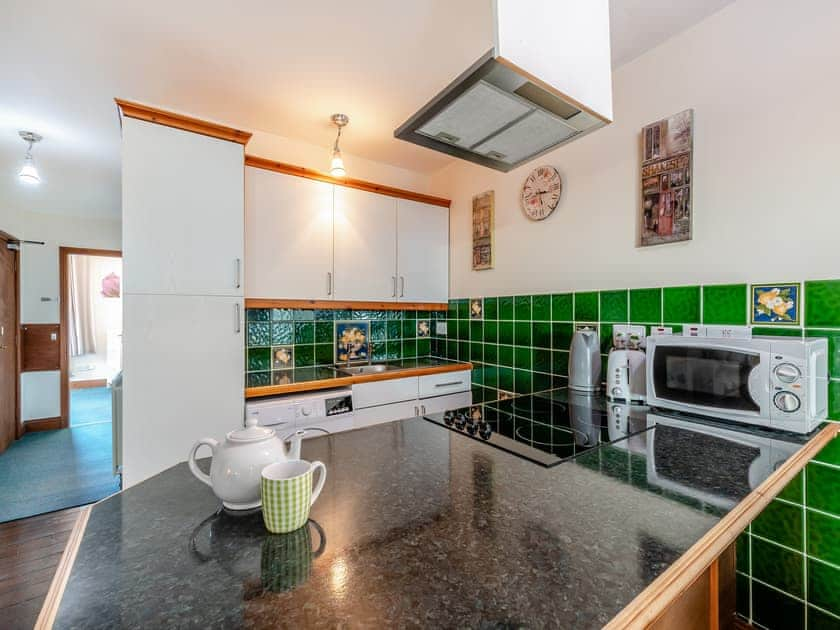 Property for sale by Auction A modern kitchen with green tiled backsplash and white cabinets in The Old Drapery Apartments at 12 Main St Haltwhistle, for sale by auction. The countertop features a teapot, a cup, and a kettle. Appliances such as a microwave, coffeemaker, and oven are visible. A clock and paintings adorn the well-lit space. Presented by Palace Auctions
