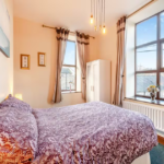 Property for sale by Auction A cozy bedroom at The Old Drapery Apartments, 12 Main St, Haltwhistle, featuring a double bed with a purple and white patterned bedspread. The room boasts two large windows with beige curtains, a white wardrobe, wall art, framed certificates, hanging lights, and a vase of flowers on the windowsill. This charming property is for sale by auction. Presented by Palace Auctions