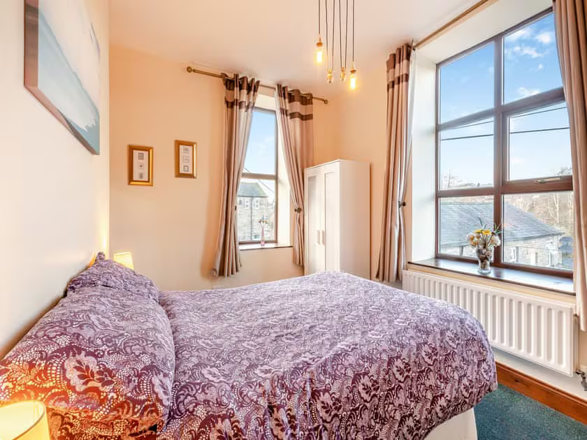 Property for sale by Auction A cozy bedroom at The Old Drapery Apartments, 12 Main St, Haltwhistle, featuring a double bed with a purple and white patterned bedspread. The room boasts two large windows with beige curtains, a white wardrobe, wall art, framed certificates, hanging lights, and a vase of flowers on the windowsill. This charming property is for sale by auction. Presented by Palace Auctions
